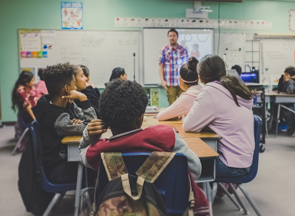 A photo of children listening to teacher in class, taken from a child's perspective.